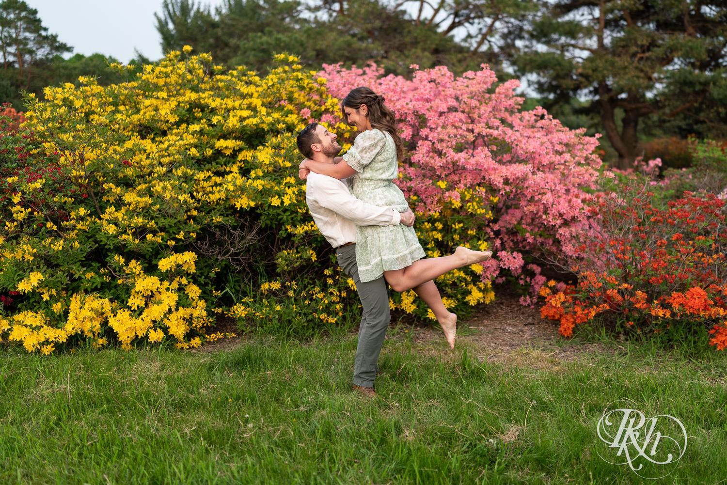 Man in white shirt lifts woman in green dress during engagement photography at the Minnesota Landscape Arboretum in Chaska, Minnesota.
