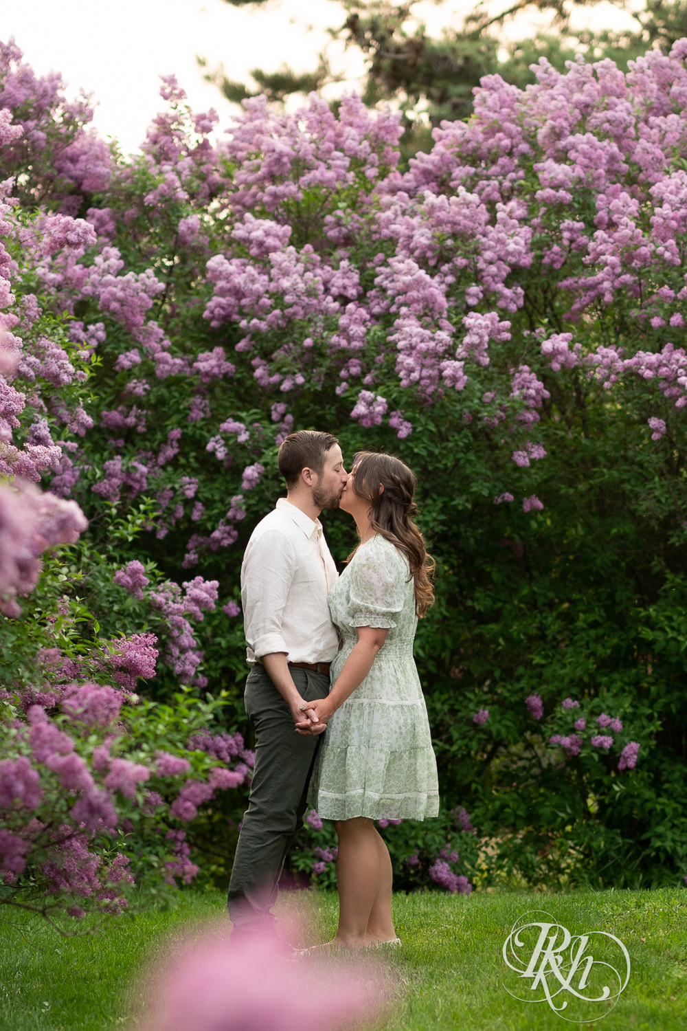 Man in white shirt and woman in green kiss in front of lilacs during engagement photography at the Minnesota Landscape Arboretum in Chaska, Minnesota.