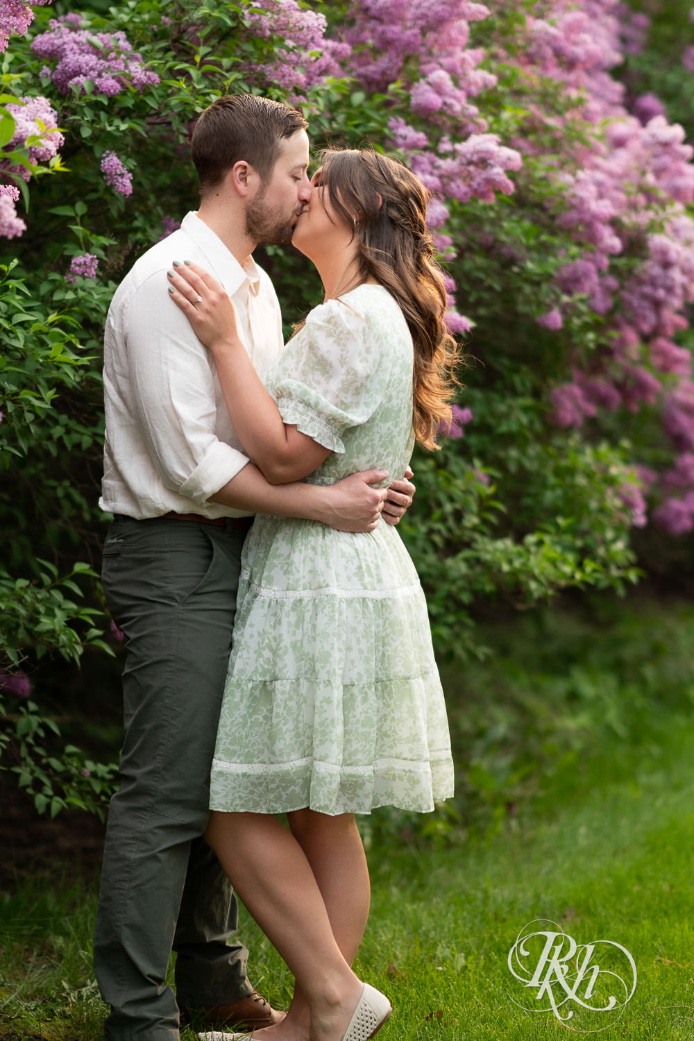 Man in white shirt and woman in green kiss in front of lilacs during engagement photography at the Minnesota Landscape Arboretum in Chaska, Minnesota.