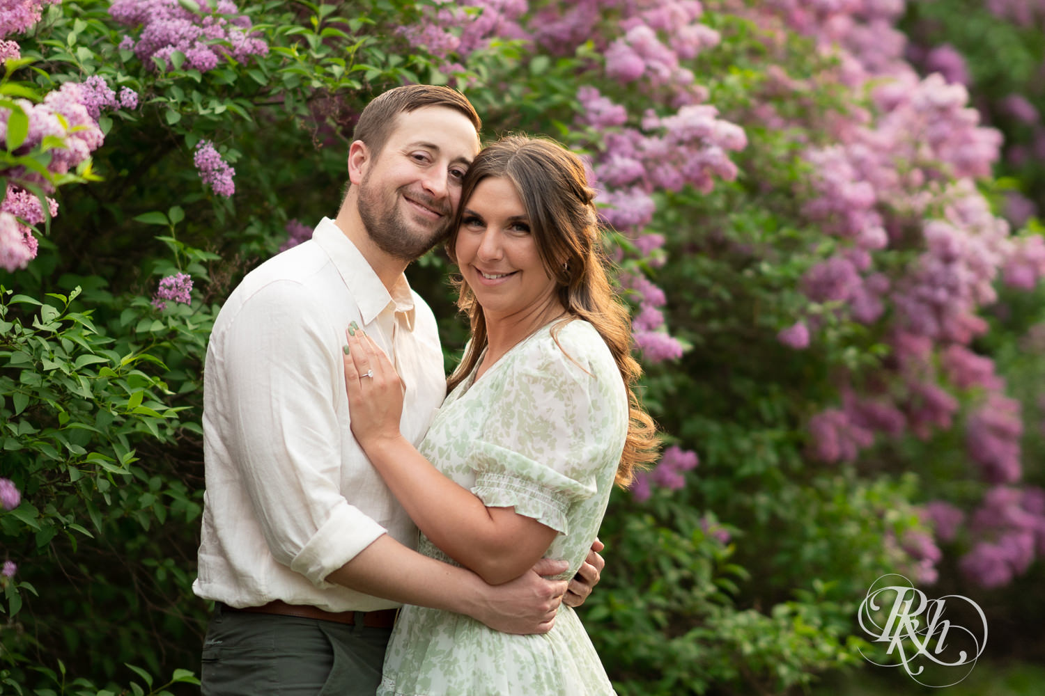 Man in white shirt and woman in green smile in front of lilacs during engagement photography at the Minnesota Landscape Arboretum in Chaska, Minnesota.