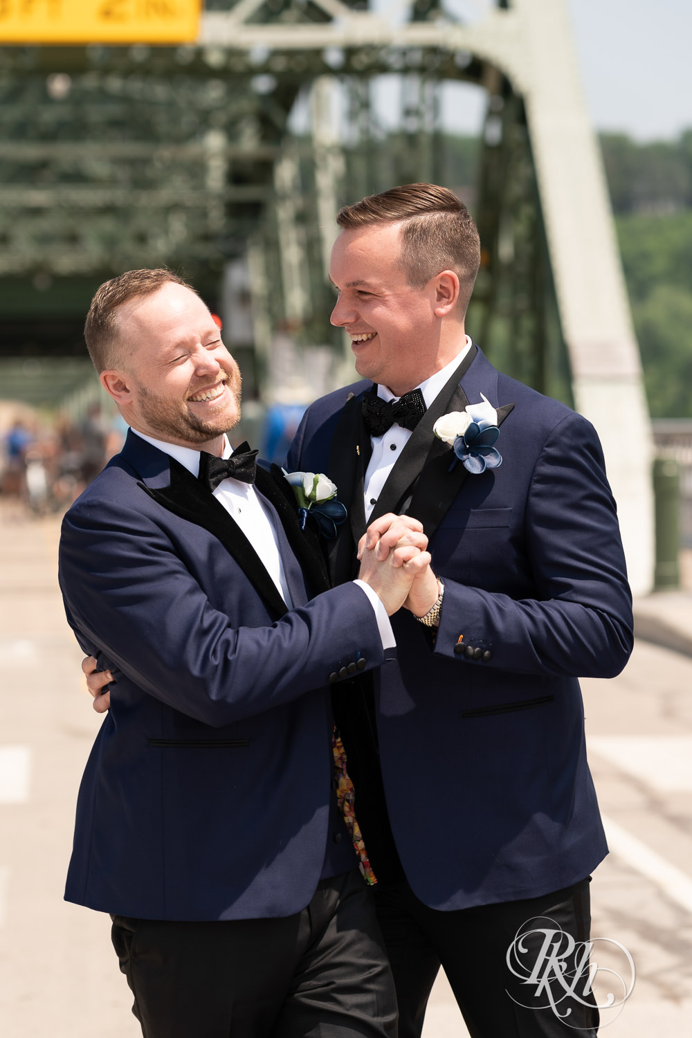 Grooms in blue tuxedos laugh on bridge before gay wedding in Stillwater, Minnesota.