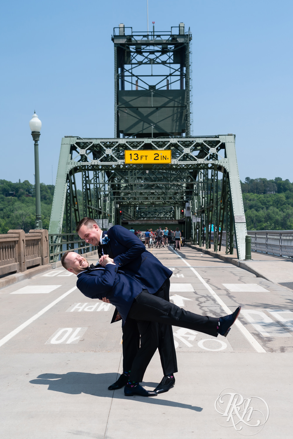Grooms in blue tuxedos laugh on bridge before gay wedding in Stillwater, Minnesota.