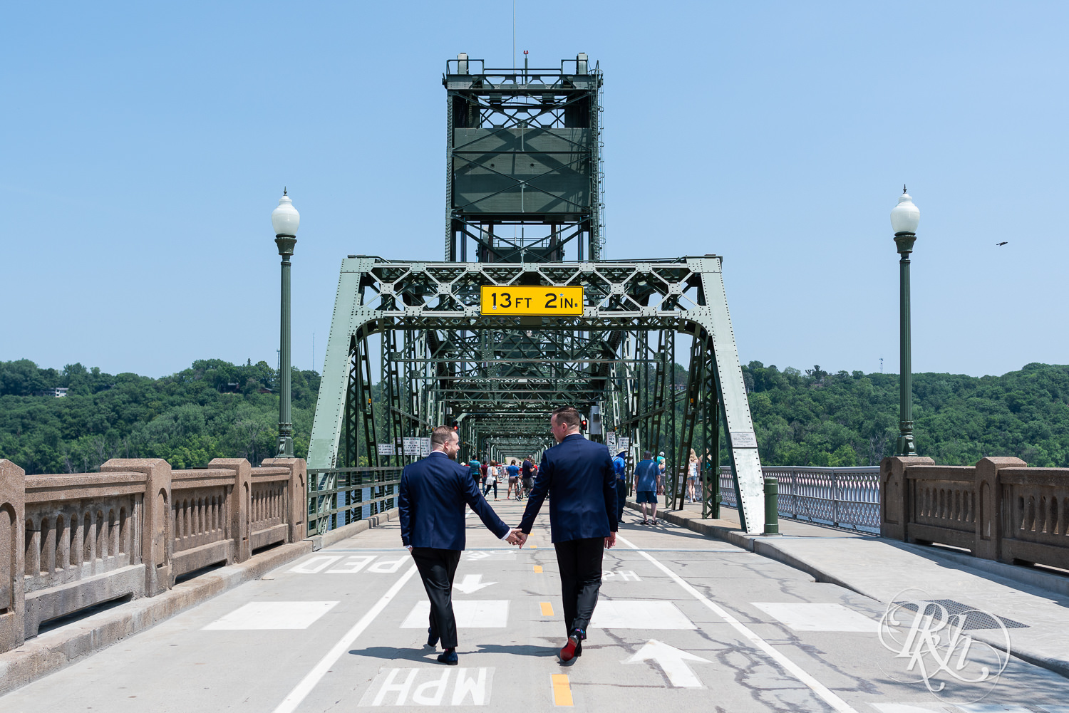 Grooms in blue tuxedos hold hands on bridge before gay wedding in Stillwater, Minnesota.