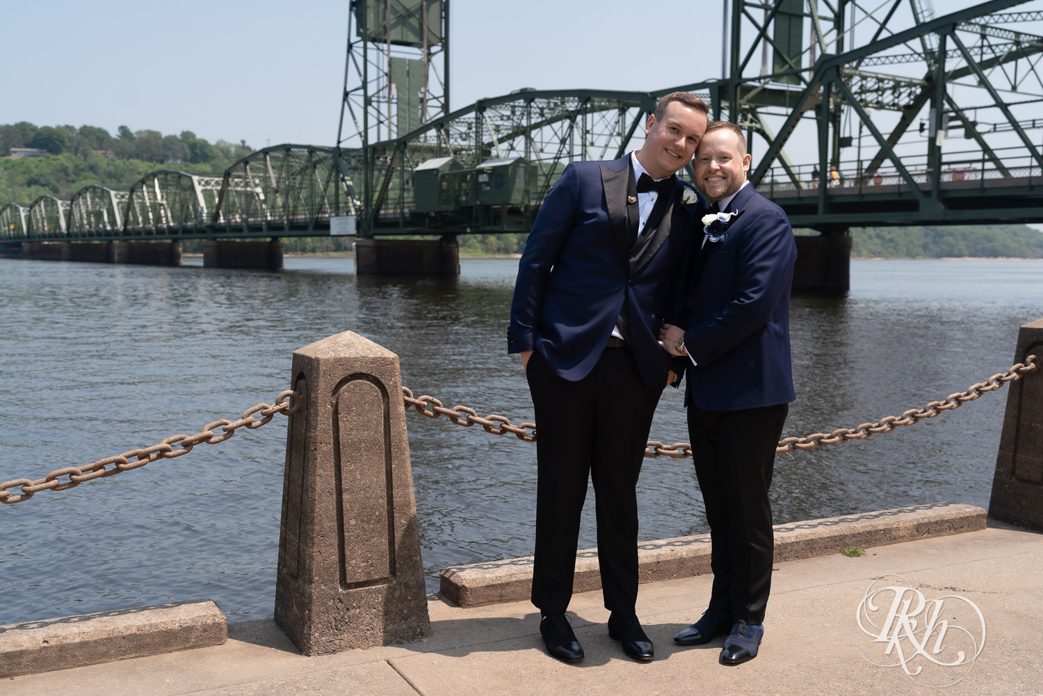 Grooms in blue tuxedos smile in front of river before gay wedding in Stillwater, Minnesota.