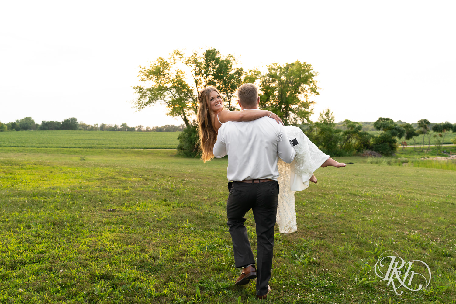 Groom carries bride across the field during sunset at Ahavah Cottage in Elysian, Minnesota.