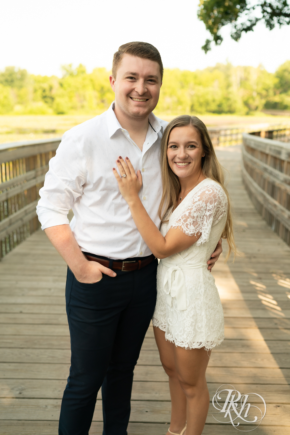Man and woman dressed in white smile on a bridge during summer engagement photography in Minnesota.