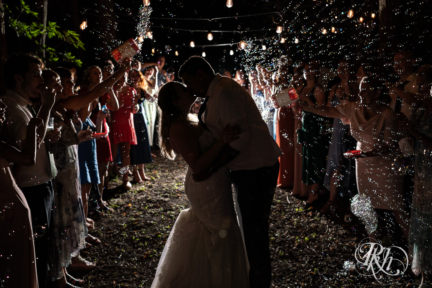 Bride and groom kiss as guests blow bubbles at them at Cottage Farmhouse in Glencoe, Minnesota.