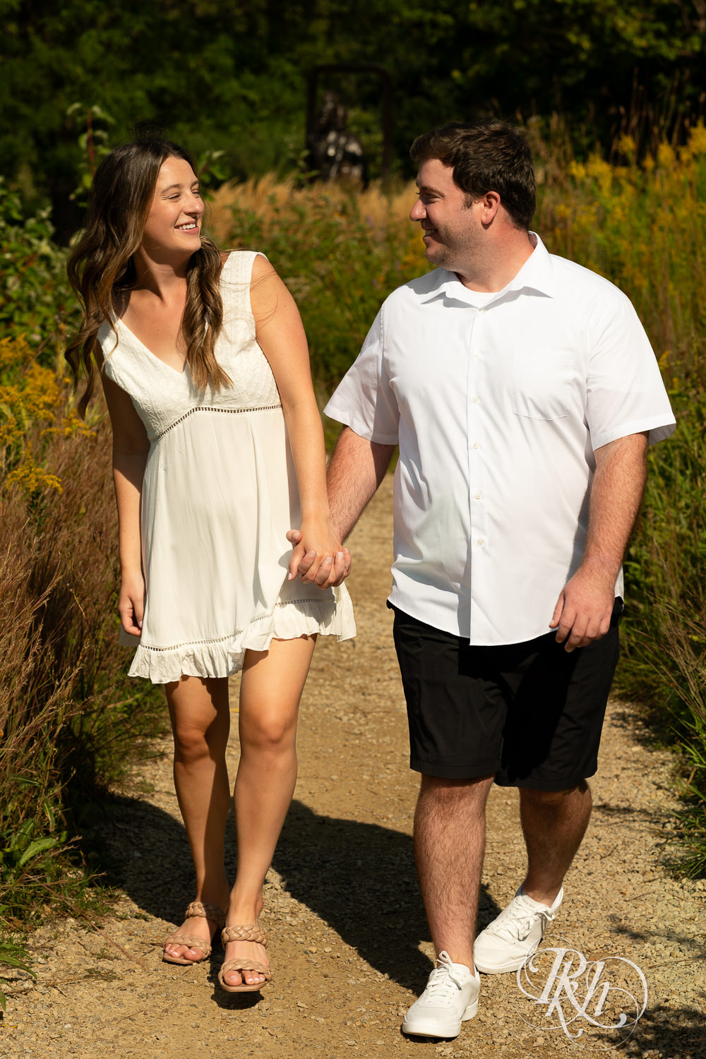 Man and woman in white dress walk together during engagement photography at Centennial Lakes Park in Edina, Minnesota.