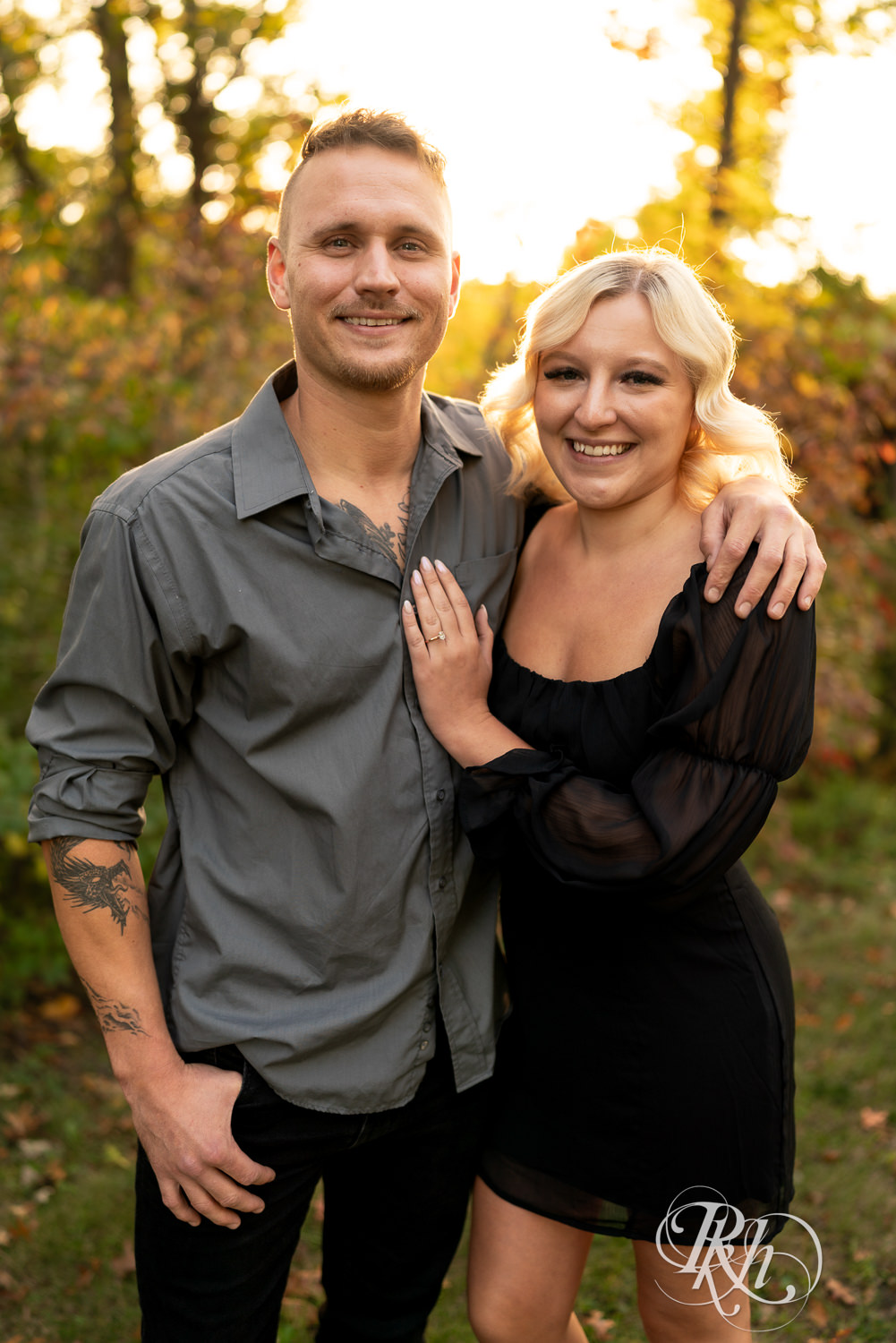 Blond woman in black dress and man in dress shirt smile during sunset engagement photos at Lebanon Hills Regional Park in Eagan, Minnesota.