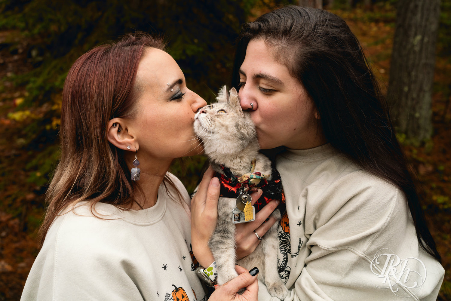 Lesbian couple in Halloween sweaters smile with their cat during fall engagement photos at Amnicon Falls in Wisconsin. 
