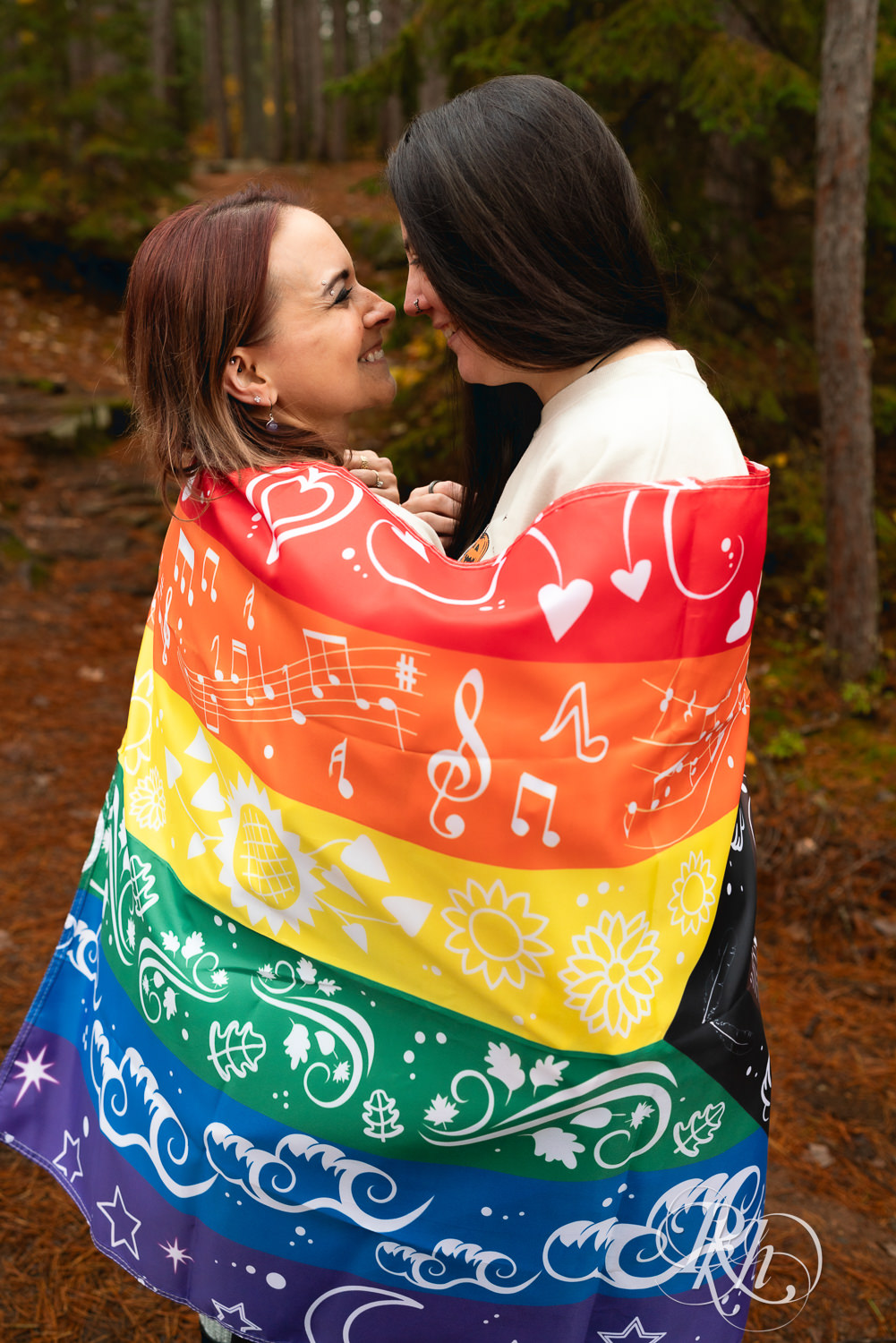 Lesbian couple smile wrapped in Pride flag during fall engagement photos at Amnicon Falls in Wisconsin. 