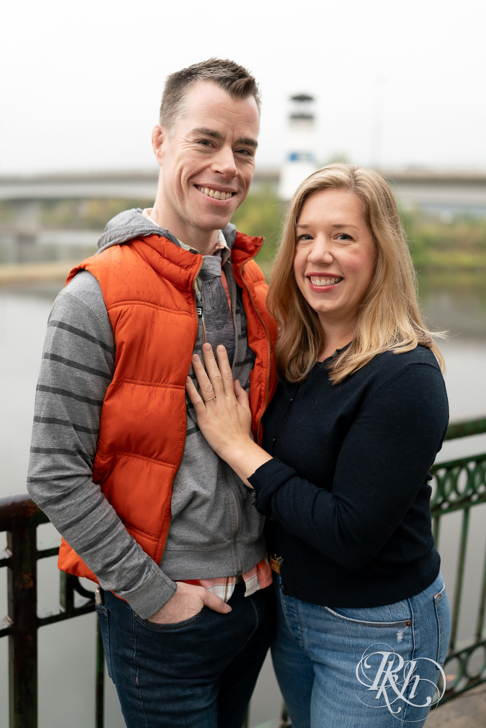 Man and woman in hoodies and jeans smile during rainy engagement photos at Boom Island Park in Minneapolis, Minnesota.