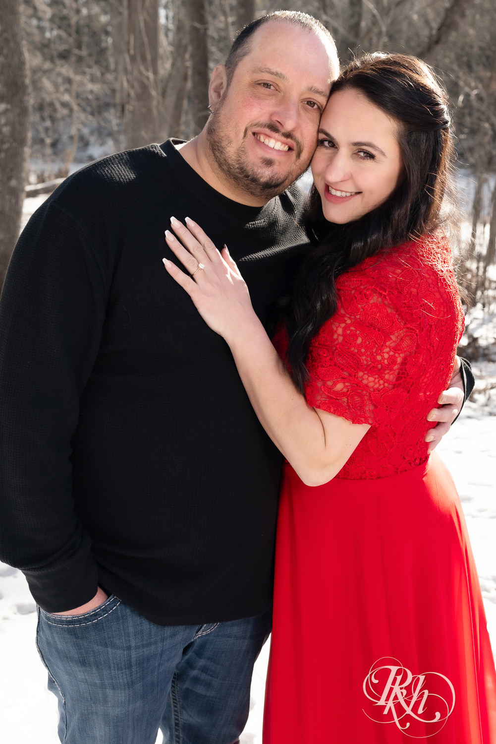 Man in jeans and woman in red dress smile in the woods during winter engagement photography in Pine City, Minnesota.