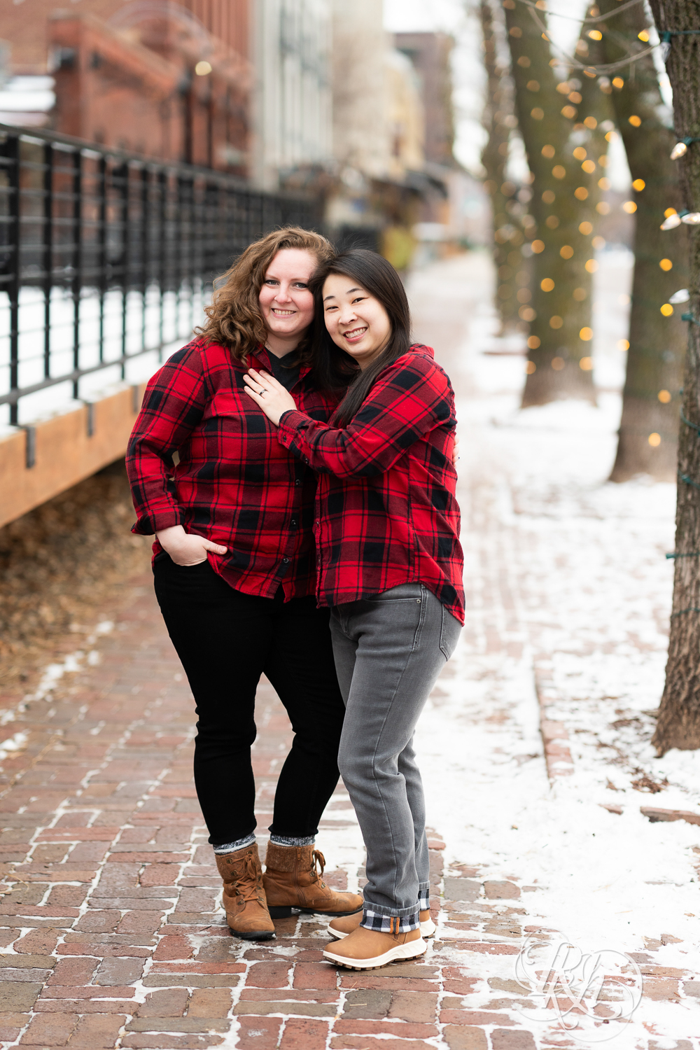 Red headed woman and Asian woman in red flannels smile during winter engagement photography in Minneapolis, Minnesota.