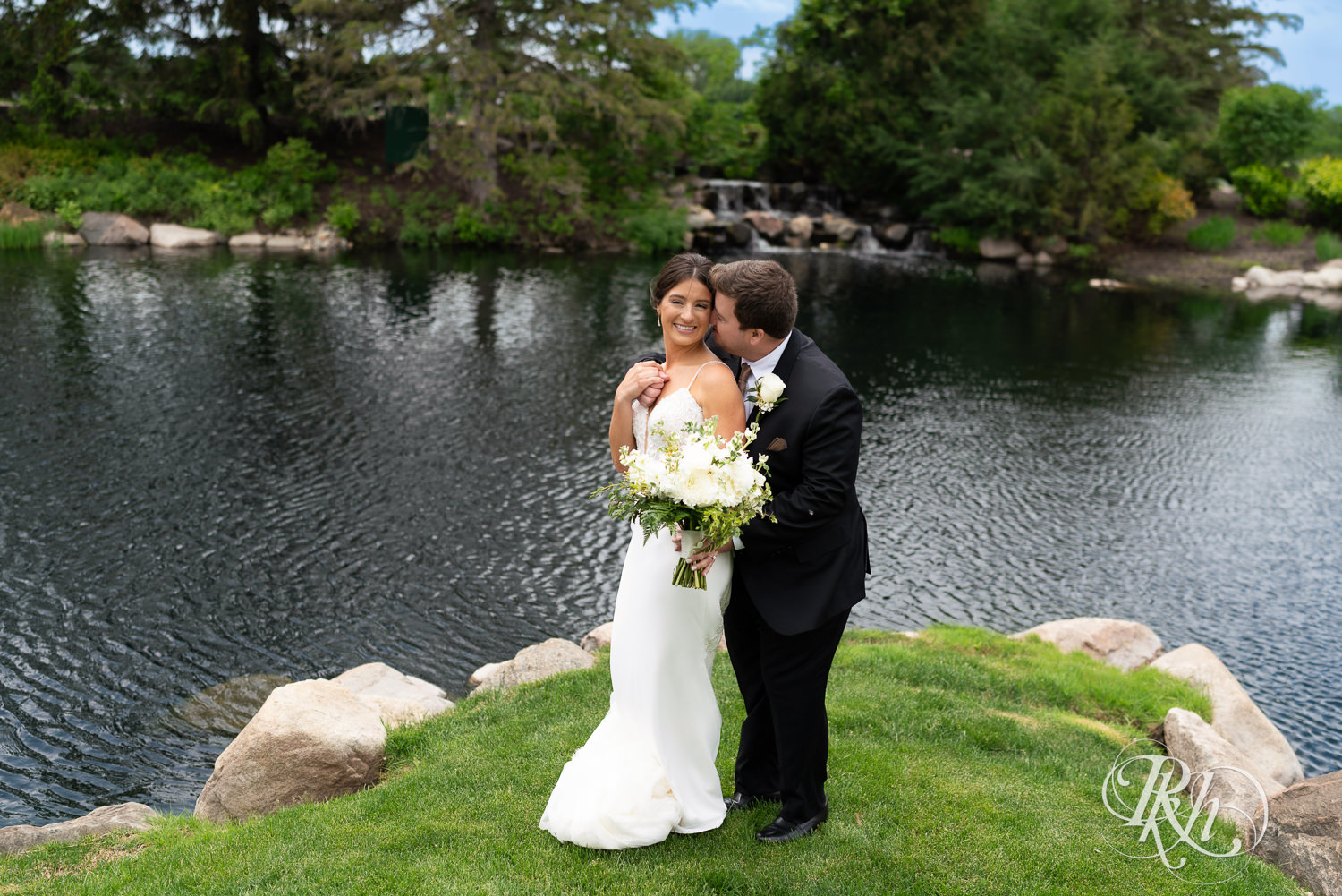 Bride and groom kiss in front of water on wedding day at Olympic Hills Golf Club in Eden Prairie, Minnesota.