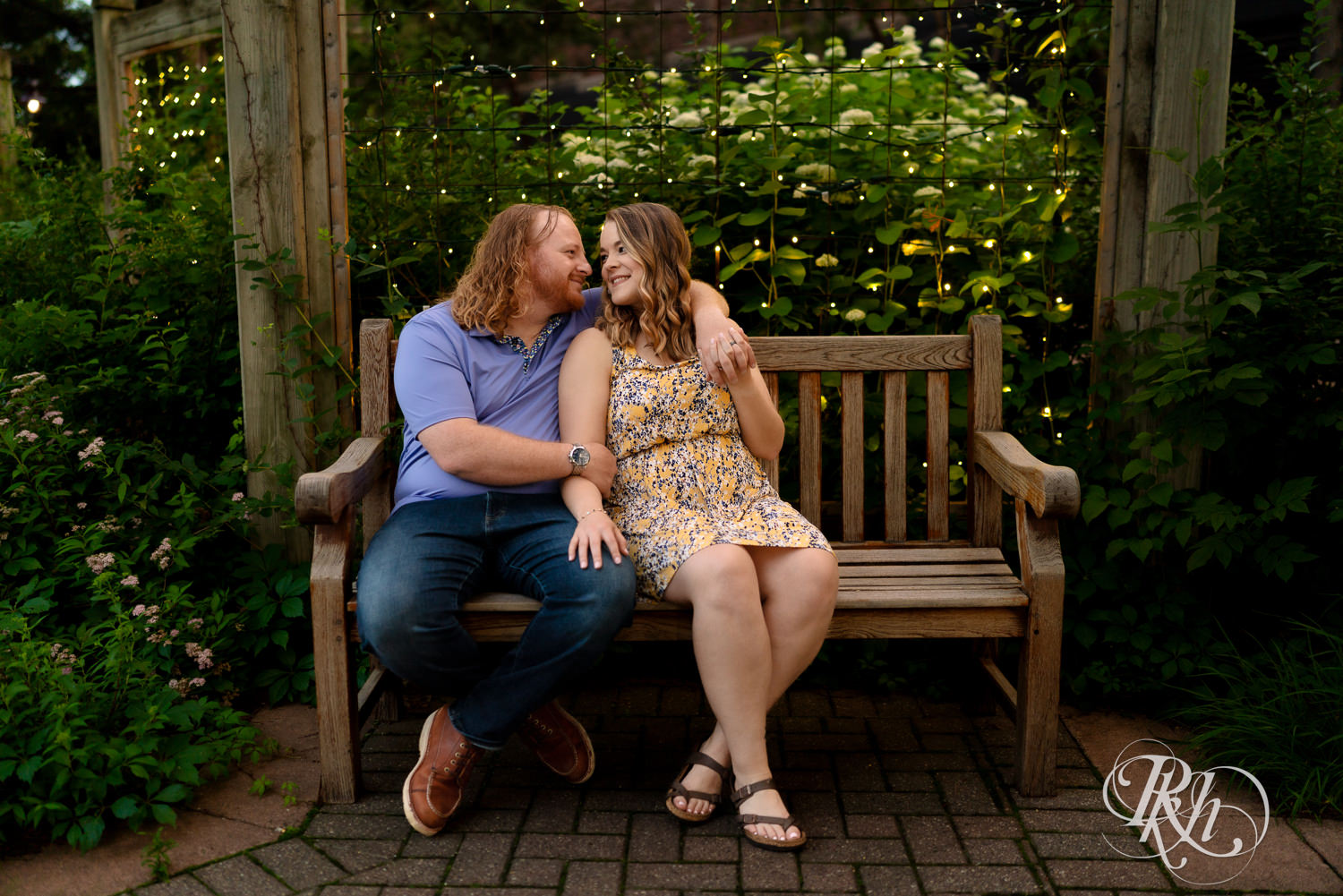 Man in blue shirt and woman in flower dress smile on a bench with a garden and string lights behind them at Centennial Lakes Park in Edina, Minnesota.