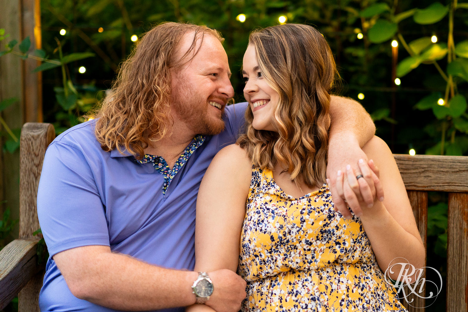 Man in blue shirt and woman in flower dress smile on a bench with a garden and string lights behind them at Centennial Lakes Park in Edina, Minnesota.