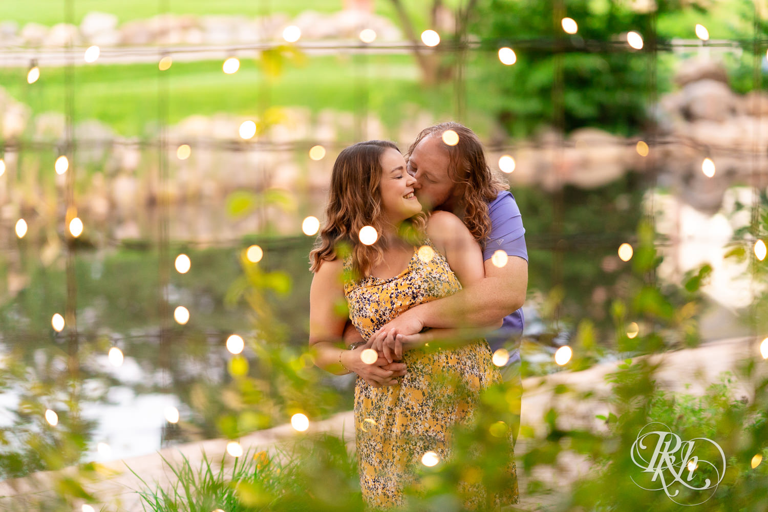 Man in blue shirt and woman in flower dress kiss behind string lights at Centennial Lakes Park in Edina, Minnesota.