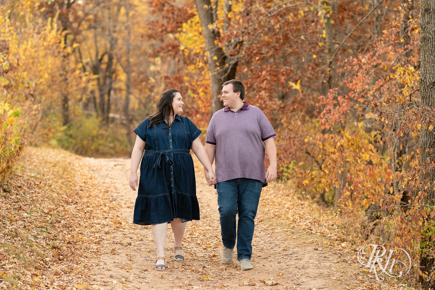 Plus size man and woman in blue dress walk in leaves during engagement photos at Lebanon Hills Regional Park in Eagan, Minnesota.