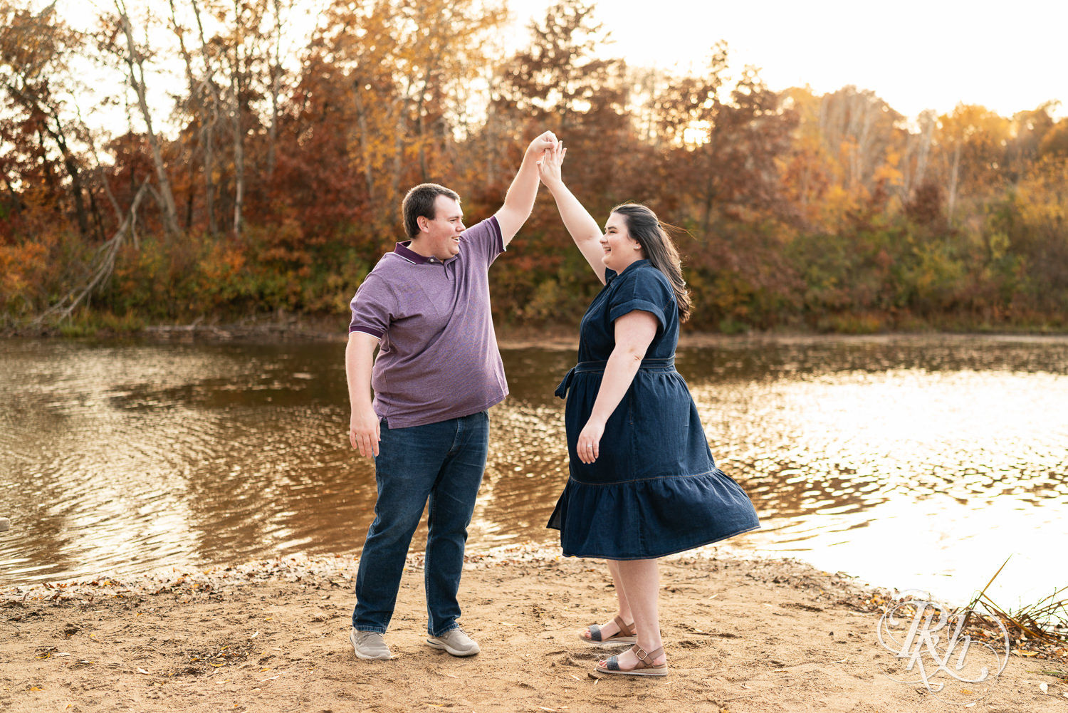 Plus size man and woman dance on beach during engagement photos at Lebanon Hills Regional Park in Eagan, Minnesota.