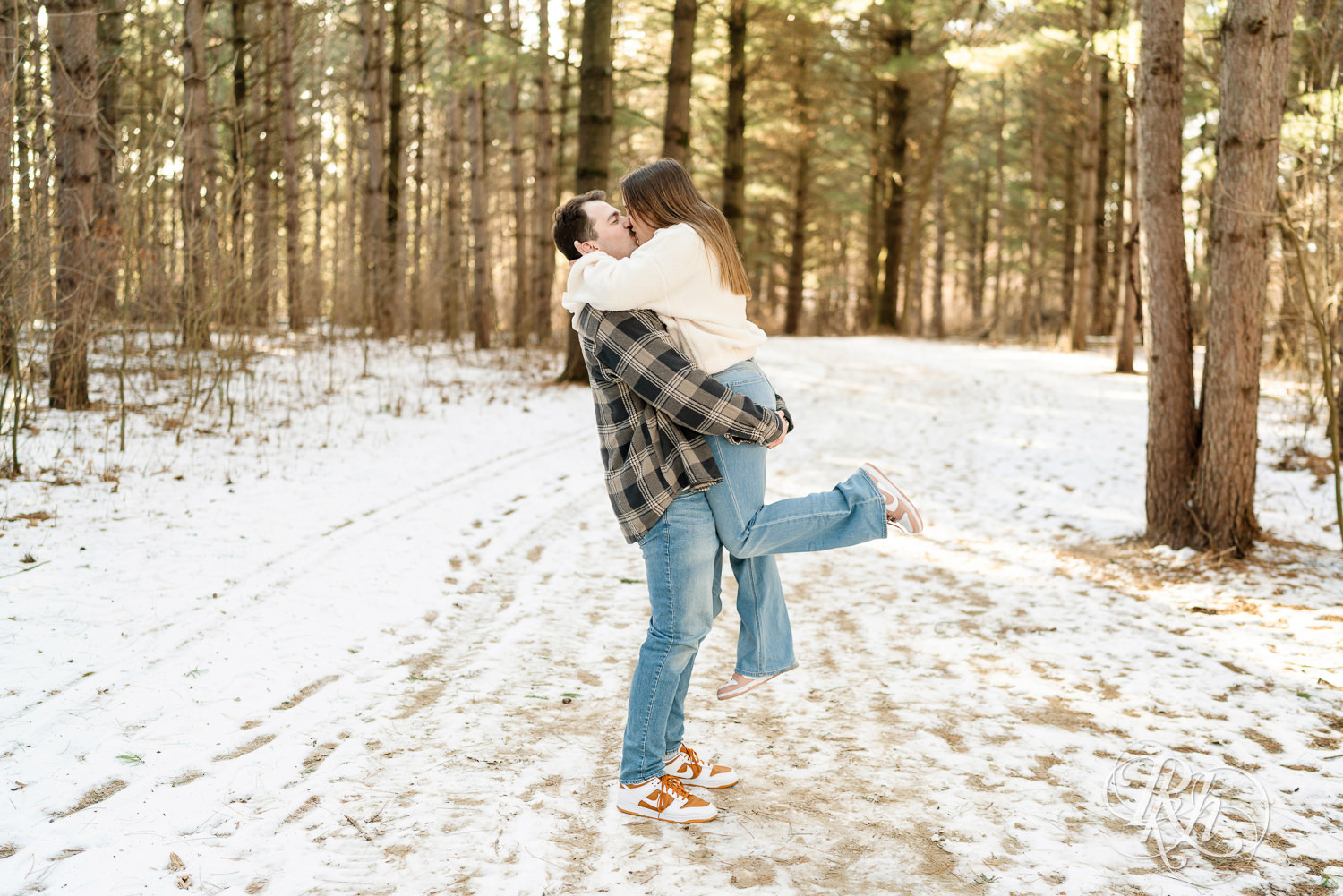 Man lifts woman in sherpa sweater in forest during winter engagement photos at Lebanon Hills Regional Park in Eagan, Minnesota.