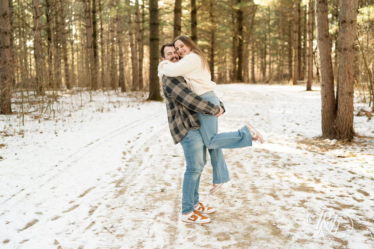 Man lifts woman in sherpa sweater in forest during winter engagement photos at Lebanon Hills Regional Park in Eagan, Minnesota.