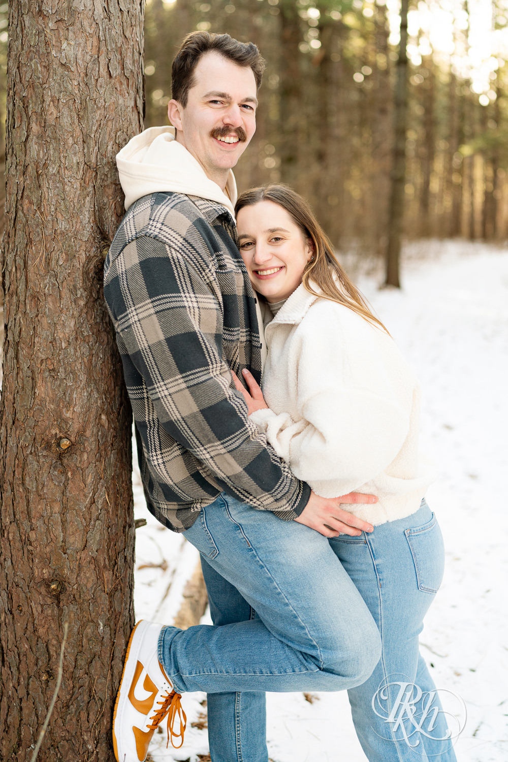 Man and woman in sherpa sweater smile in forest during winter engagement photos at Lebanon Hills Regional Park in Eagan, Minnesota.