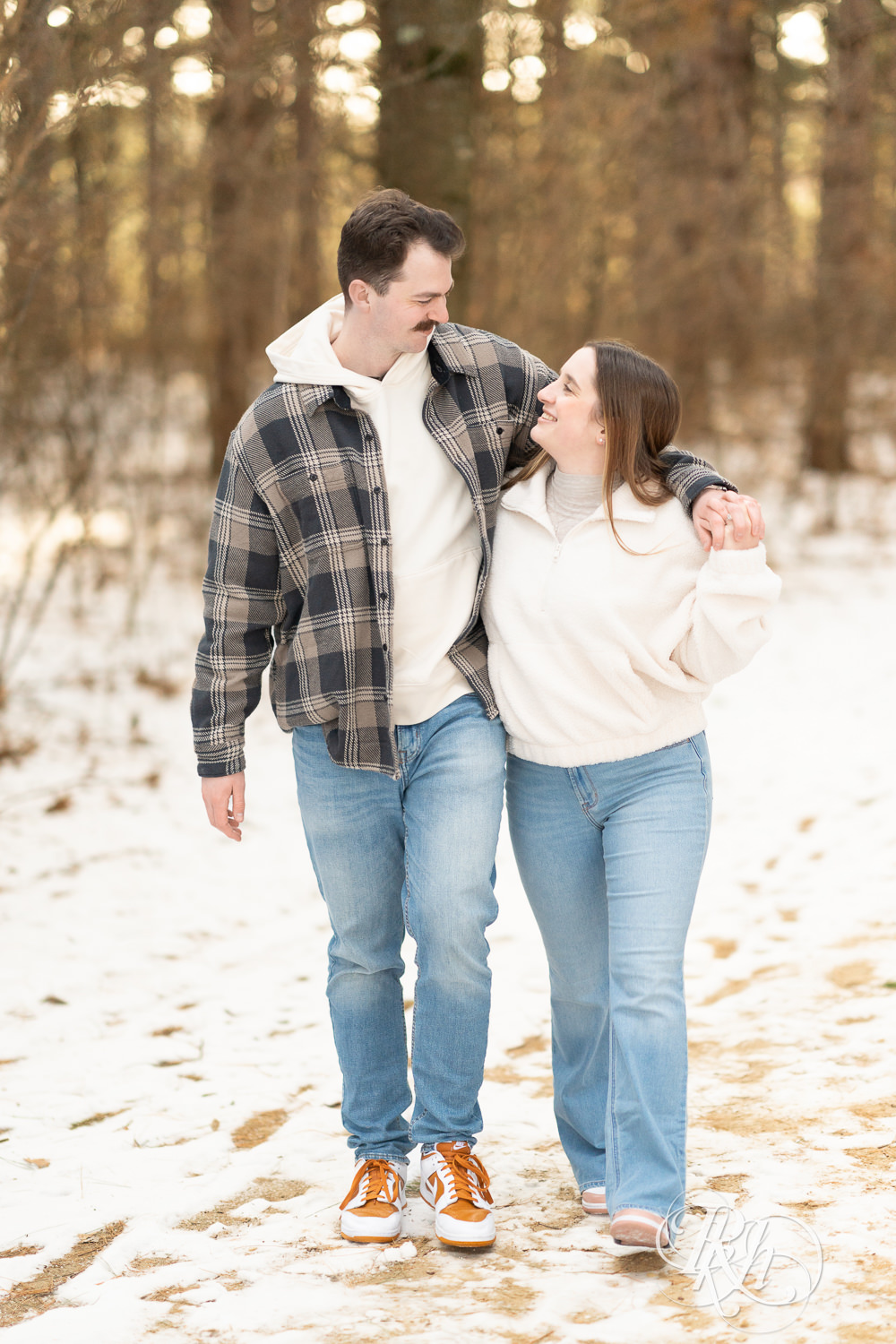 Man and woman in sherpa sweater walk in forest during winter engagement photos at Lebanon Hills Regional Park in Eagan, Minnesota.