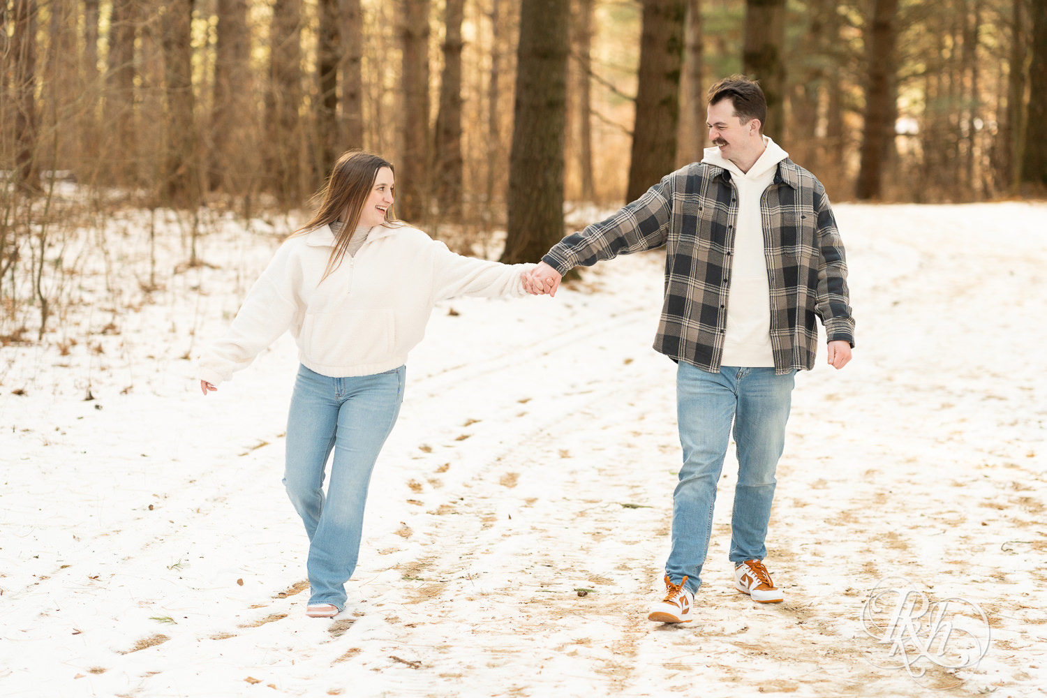 Man and woman in sherpa sweater walk in forest during winter engagement photos at Lebanon Hills Regional Park in Eagan, Minnesota.