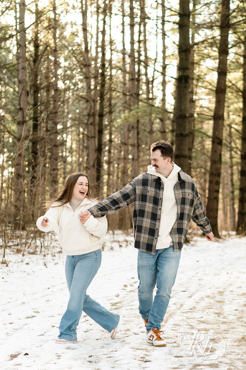 Man and woman in sherpa sweater walk in forest during winter engagement photos at Lebanon Hills Regional Park in Eagan, Minnesota.