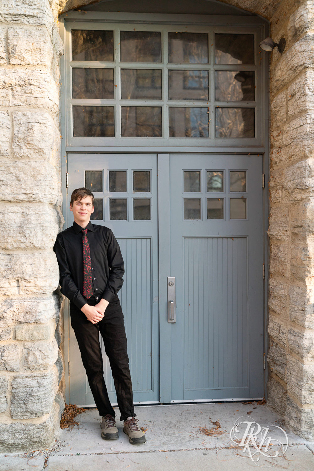 Senior boy in black dress shirt and jeans leans on brick wall in Saint Anthony Main in Minneapolis, Minnesota.
