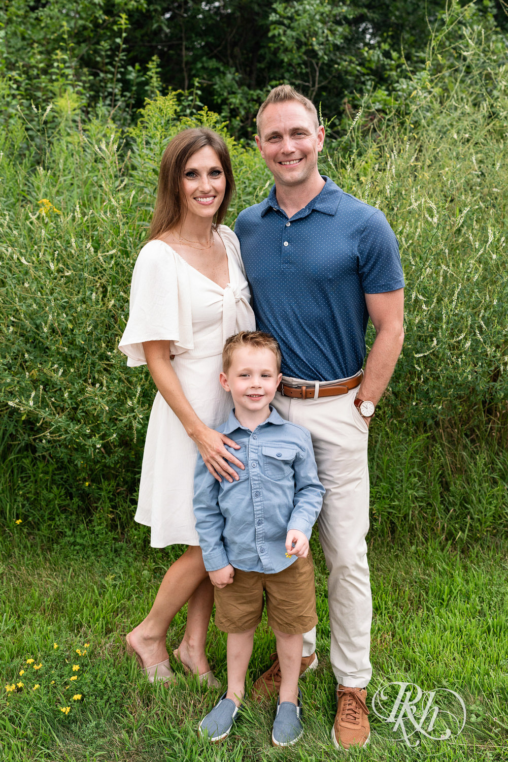 Woman in white dress and man in blue shirt smile with their son dressed in blue in field in Eagan, Minnesota.