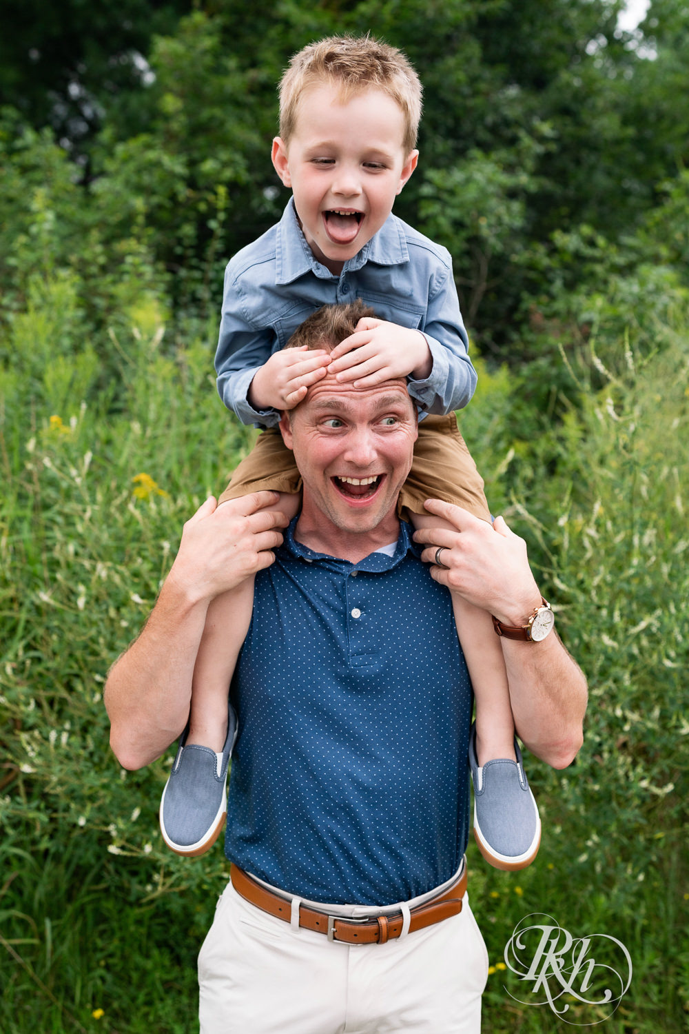 Boy dressed in blue sits on his dad's shoulders laughing in field in Eagan, Minnesota.