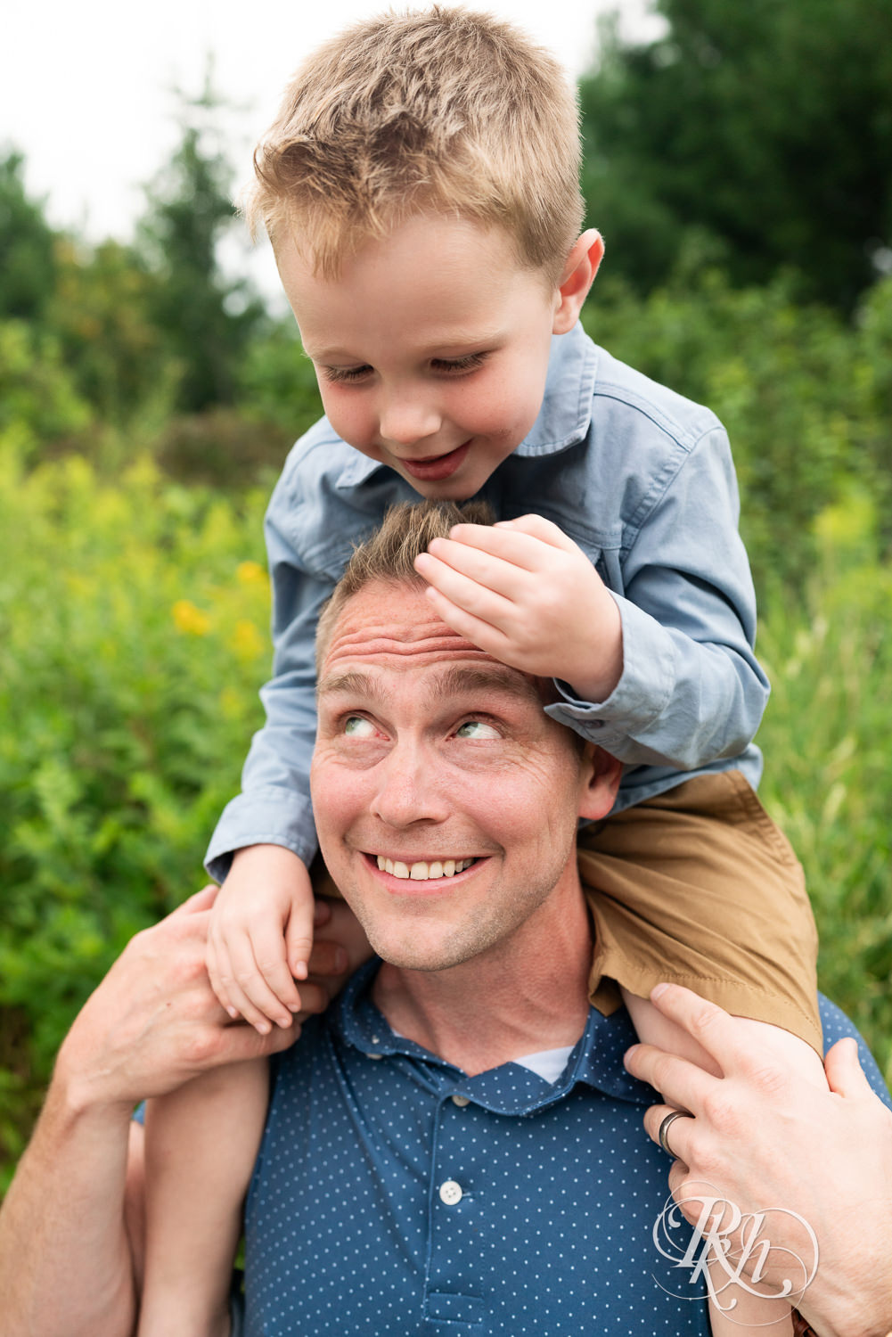 Boy dressed in blue sits on his dad's shoulders laughing in field in Eagan, Minnesota.