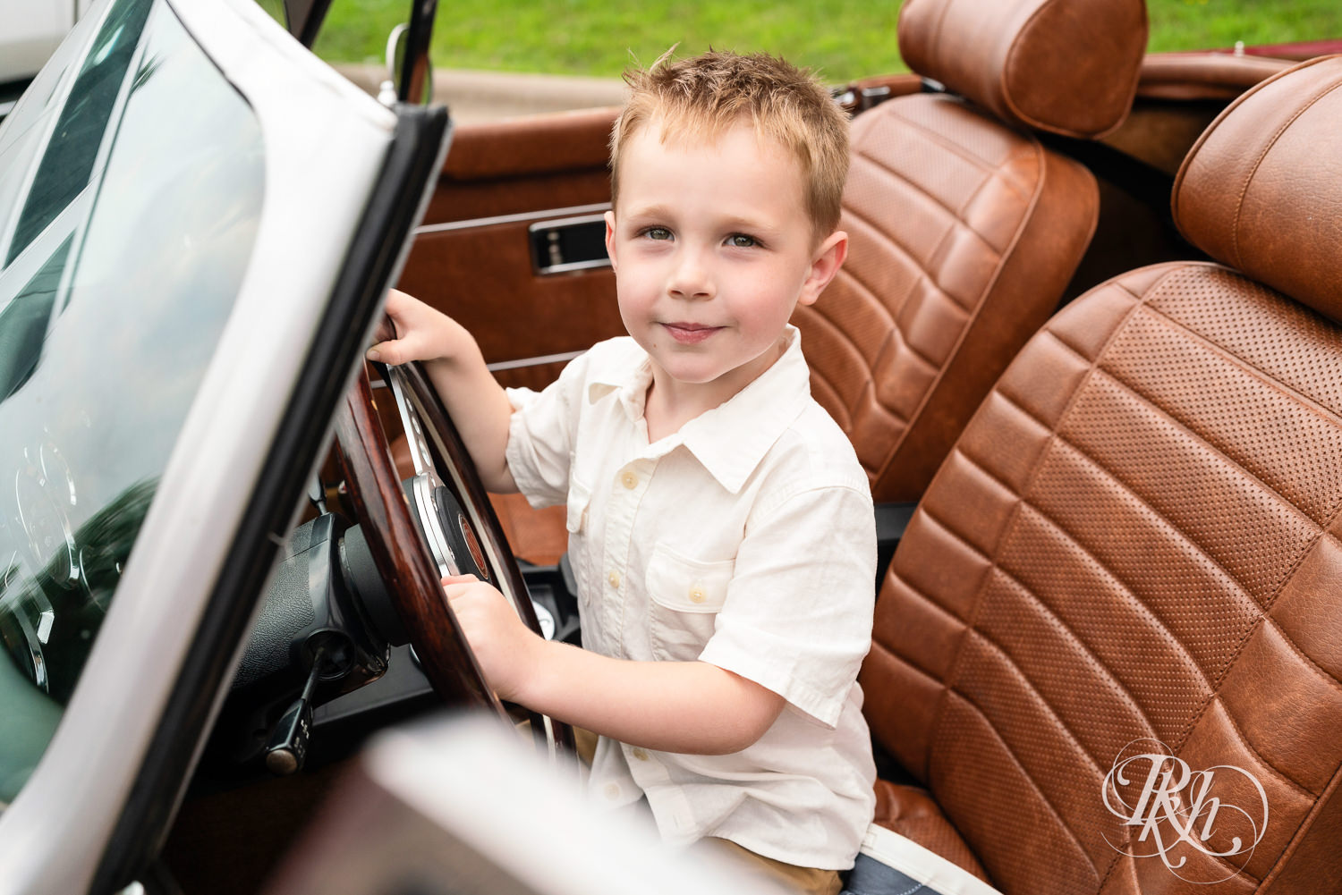 Blond boy in white dress shirt smiles in the driver's seat of a classic read MG convertible in Eagan, Minnesota.