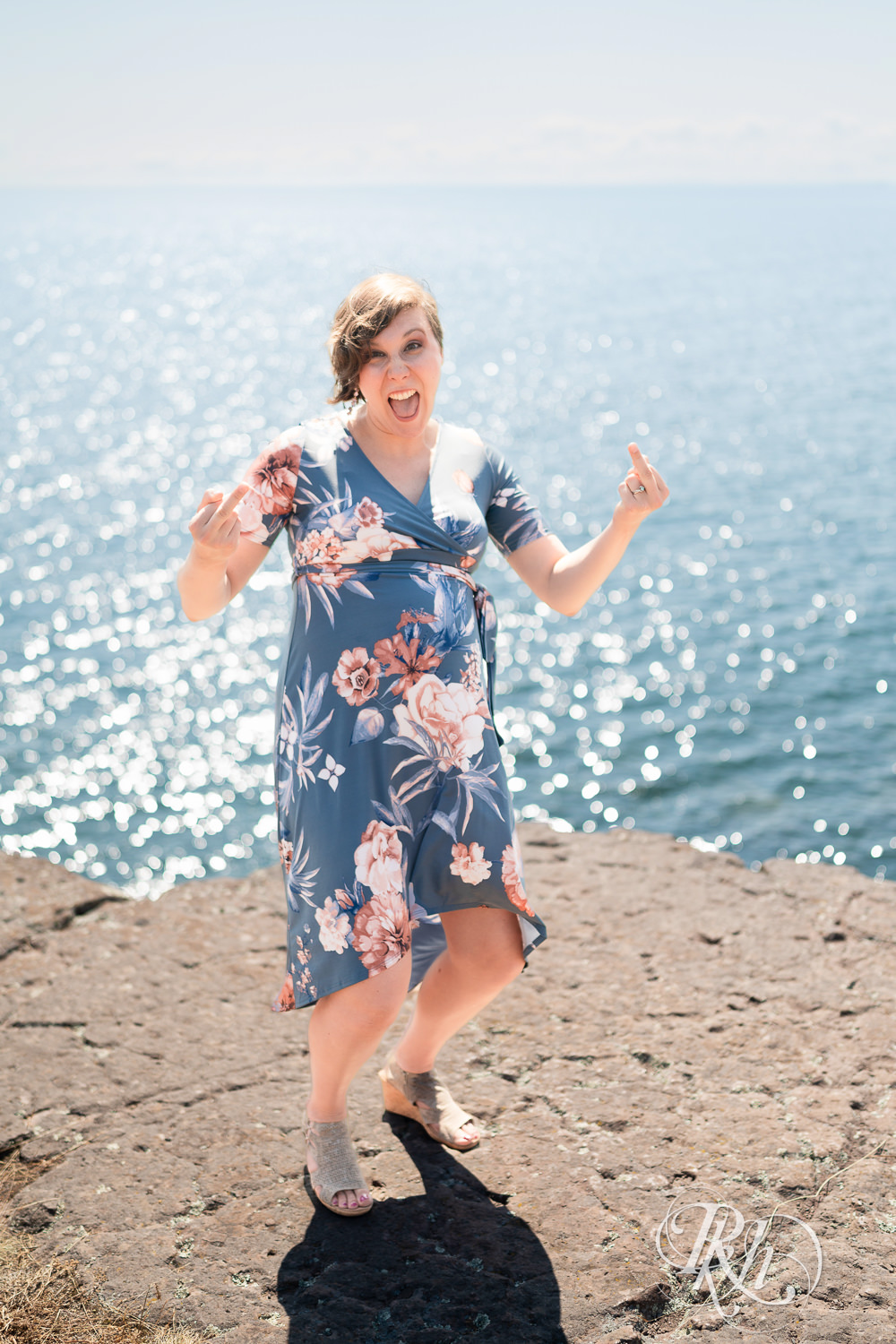 Pregnant woman with short hair in dress laughs on cliff during maternity photos in Two Harbors, Minnesota.