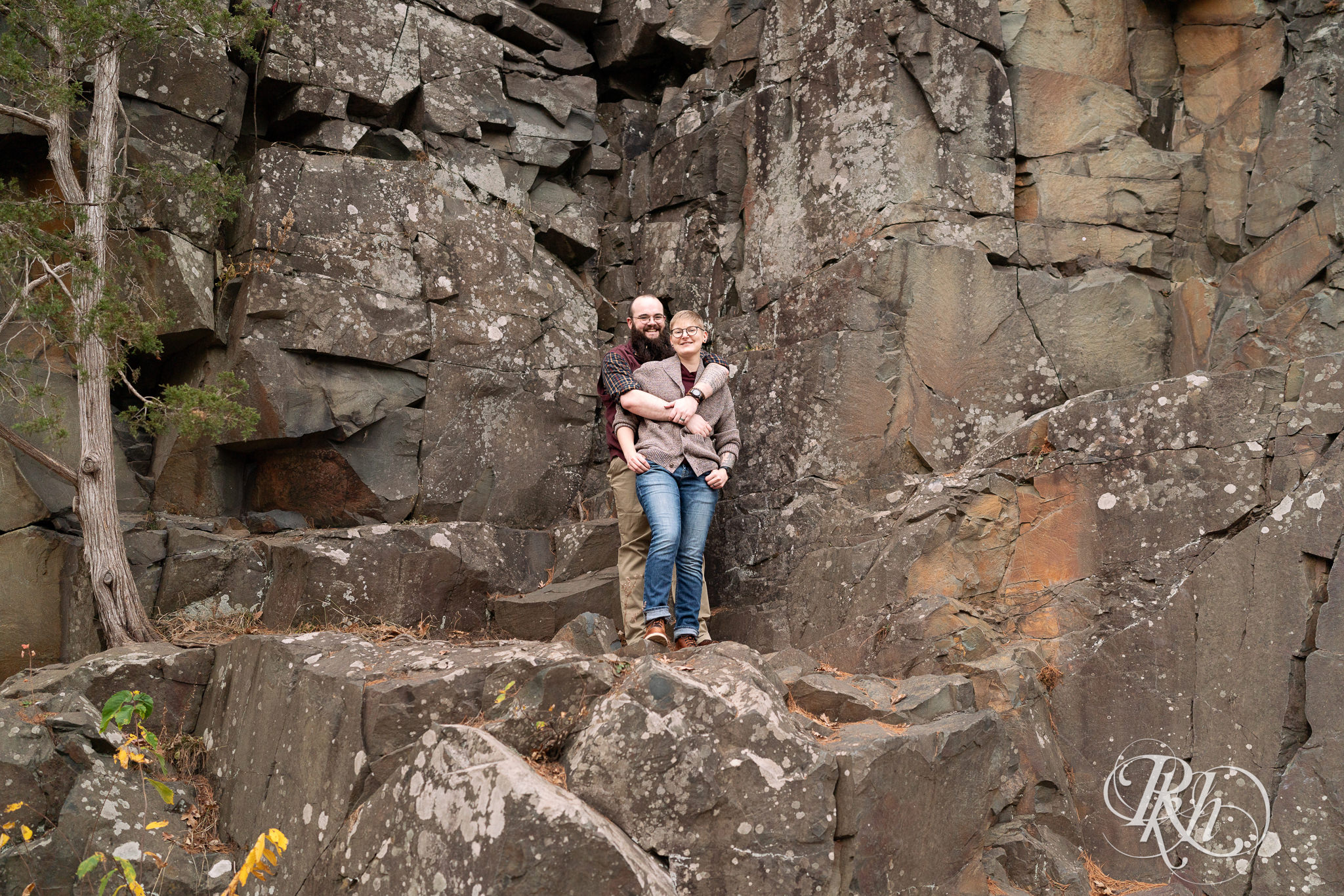 Man and non binary person smile on cliff during engagement photos in Taylor's Falls, Minnesota.