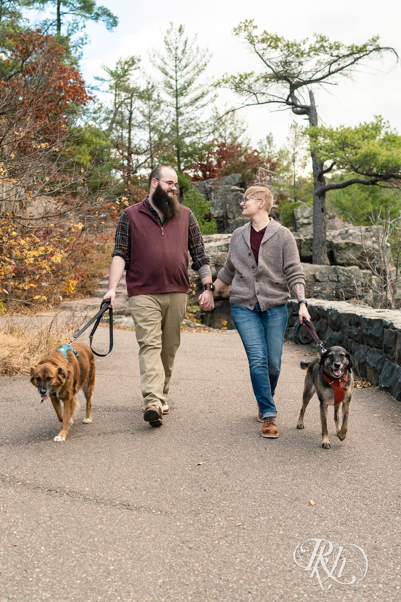 Man and non binary person walk along river with their dogs during engagement photos in Taylor's Falls, Minnesota.