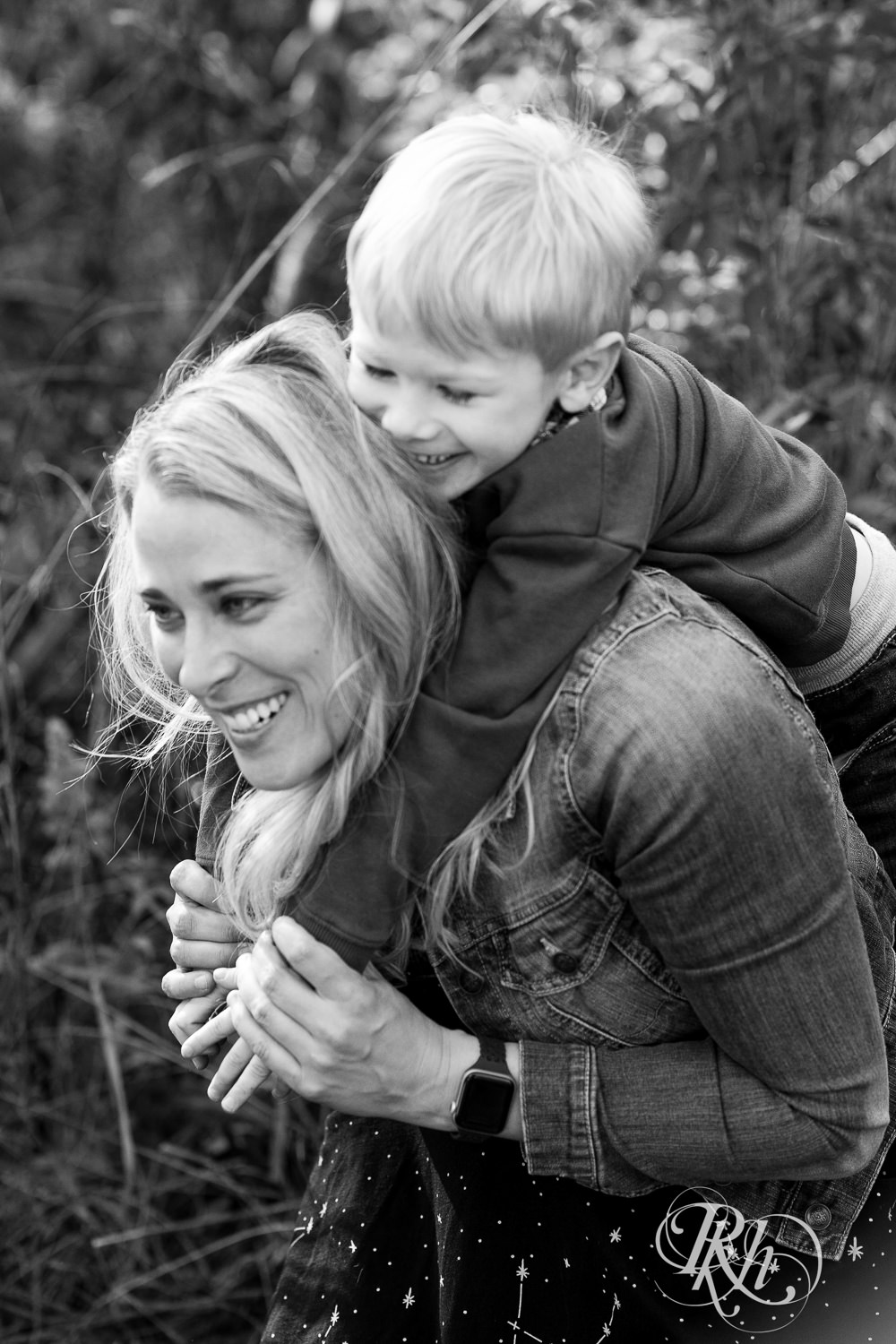 Little blond boy smiles on mom's back in Lebanon Hills Regional Park in Eagan, Minnesota.