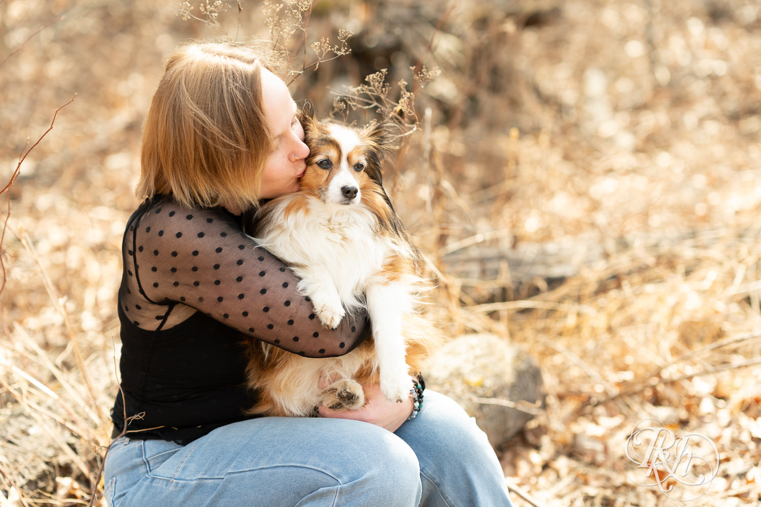 Blond woman kisses her mini Aussie dog while sitting on a log in Eagan, Minnesota during dog photography session.