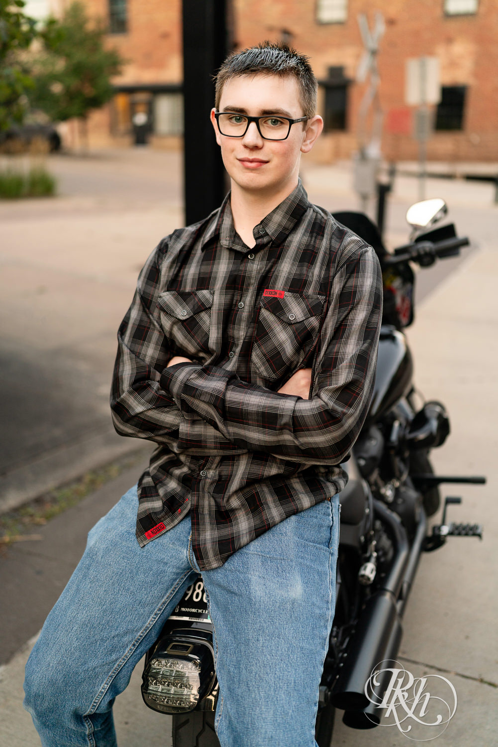 Senior boy wearing glasses and a flannel smiles sitting on a Harley Davidson motorcycle in Minneapolis, Minnesota.