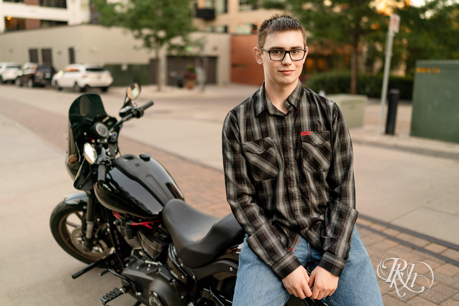 Senior boy wearing glasses and a flannel smiles sitting on a Harley Davidson motorcycle in Minneapolis, Minnesota.