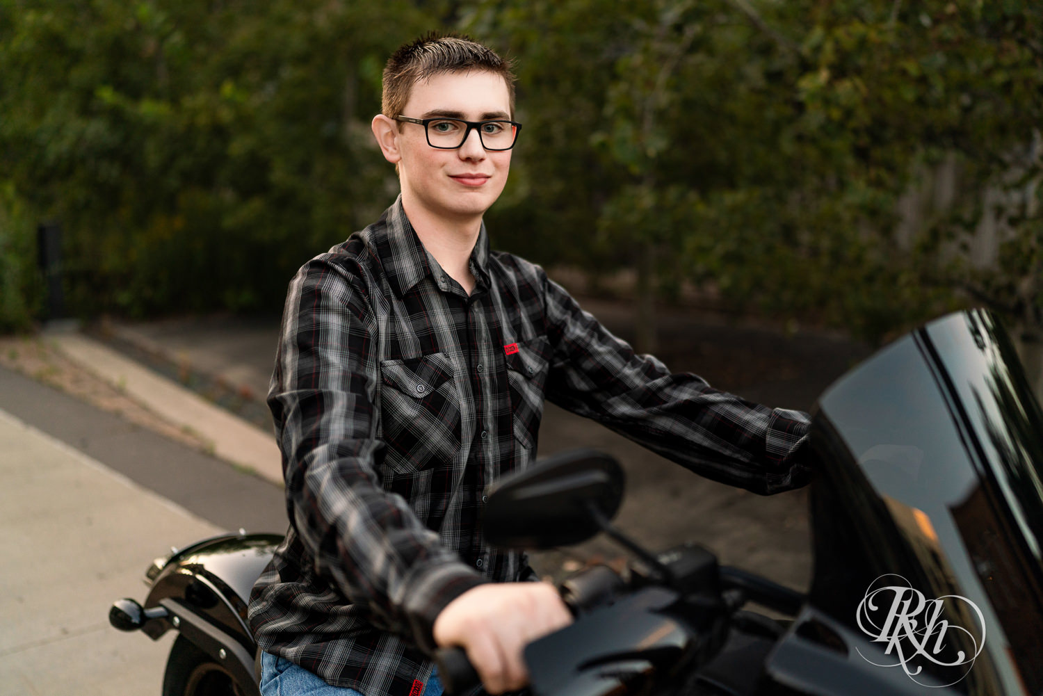 Senior boy wearing glasses and a flannel smiles sitting on a Harley Davidson motorcycle in Minneapolis, Minnesota.