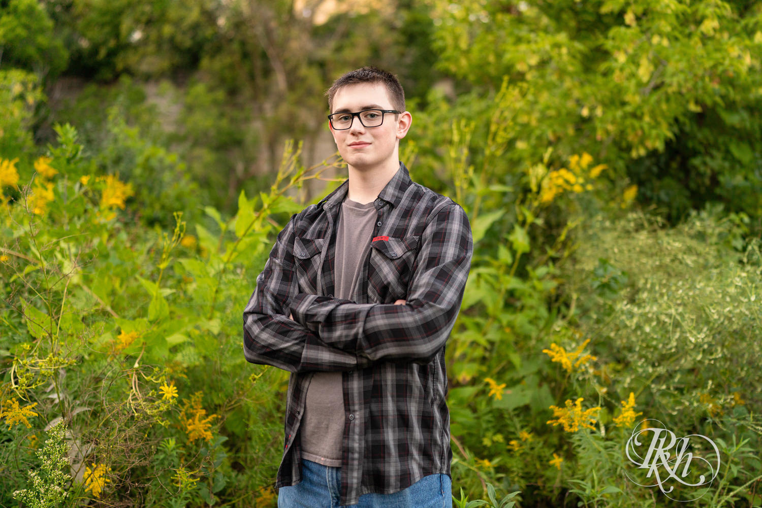 Senior boy wearing glasses and a flannel smiles in a field during sunset in Minneapolis, Minnesota.