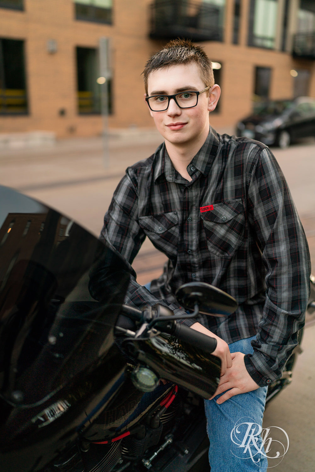 Senior boy wearing glasses and a flannel smiles sitting on a motorcycle in Minneapolis, Minnesota.