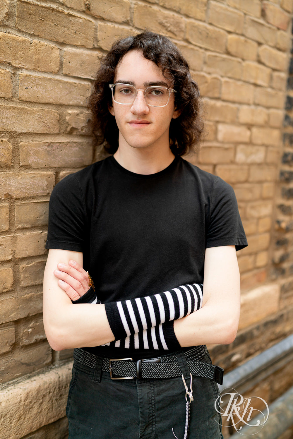 Senior boy in glasses dressed in black hoodie and jeans leans against a brick wall in Minneapolis, Minnesota.
