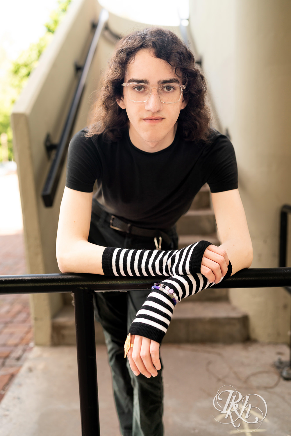 Senior boy in glasses dressed in black t-shirt and jeans leans against a railing in Minneapolis, Minnesota.