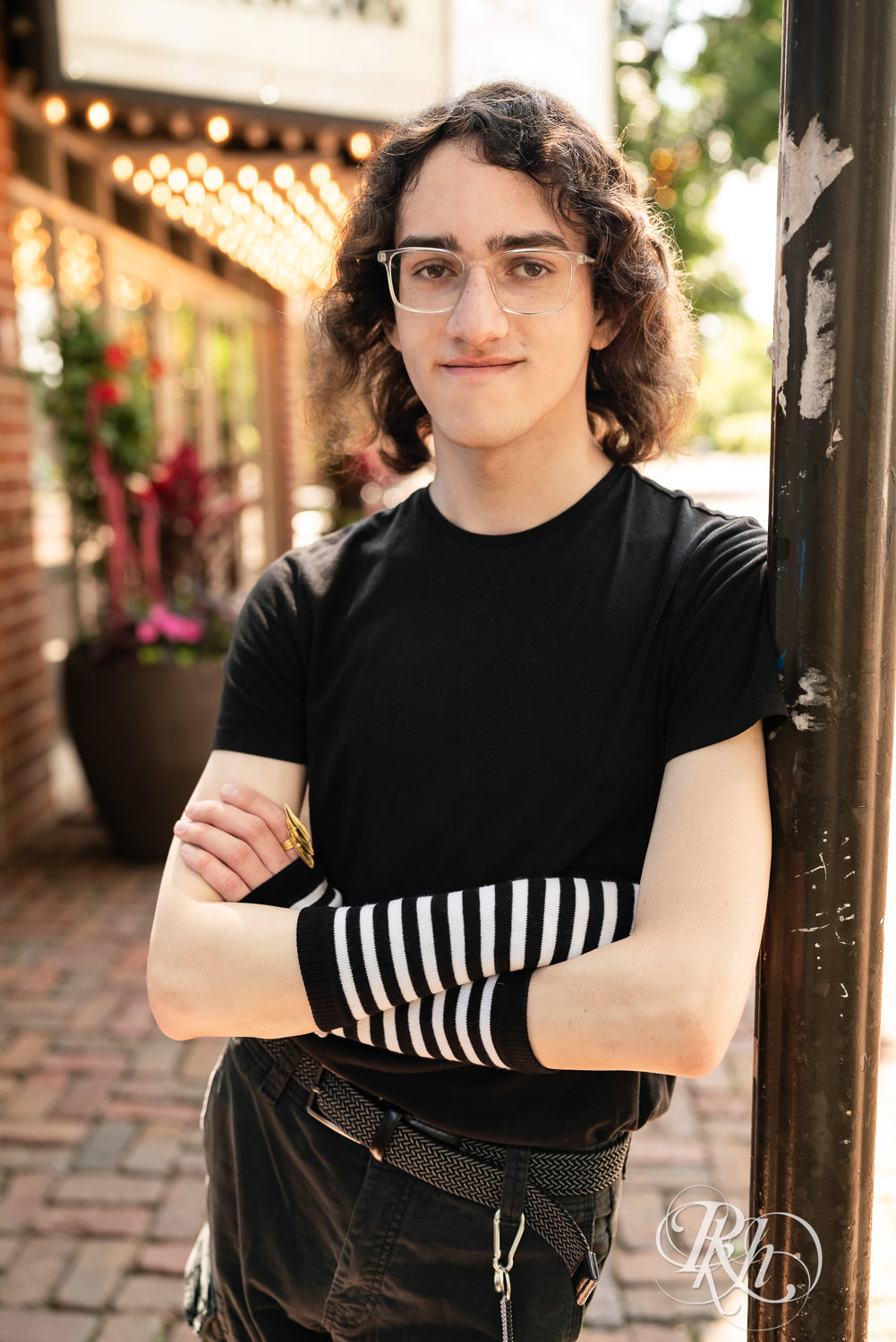 Senior boy in glasses dressed in black t-shirt and jeans leans against a pole in Minneapolis, Minnesota.