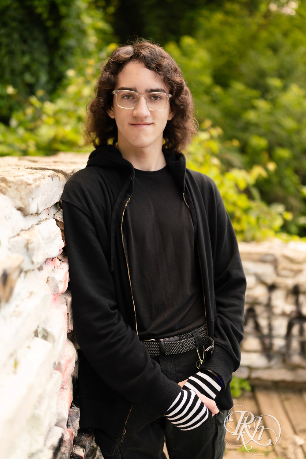 Senior boy in glasses dressed in black hoodie and jeans smiles in the lower trail in Minneapolis, Minnesota.