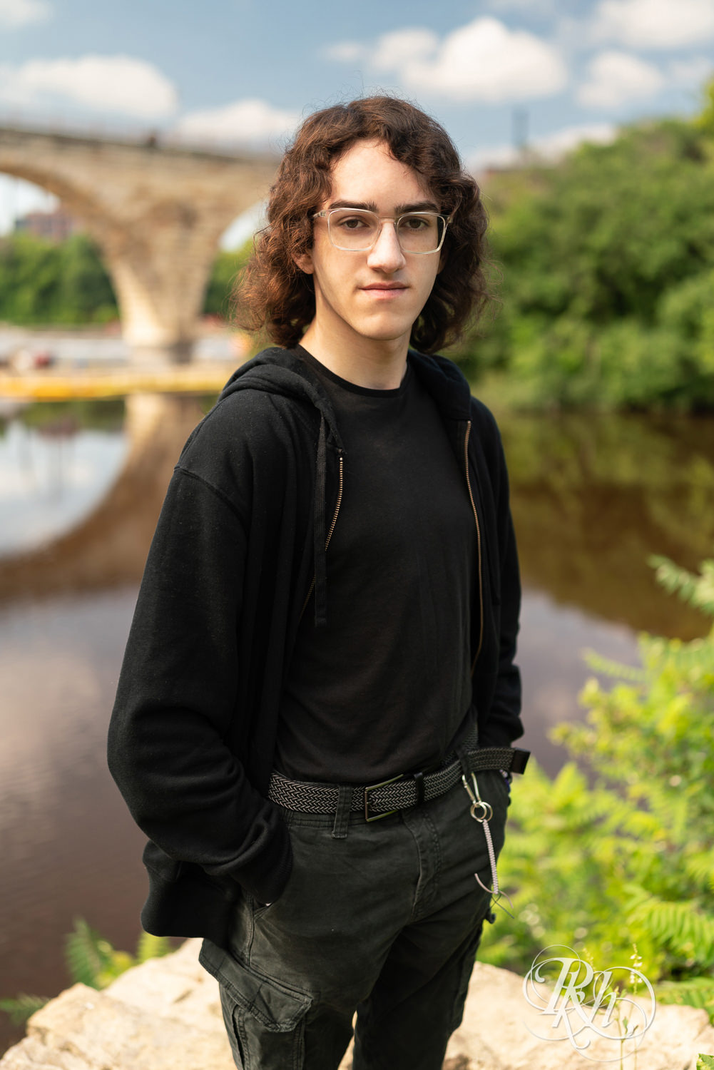 Senior boy in glasses dressed in black hoodie and jeans smiles in front of the river in Minneapolis, Minnesota.